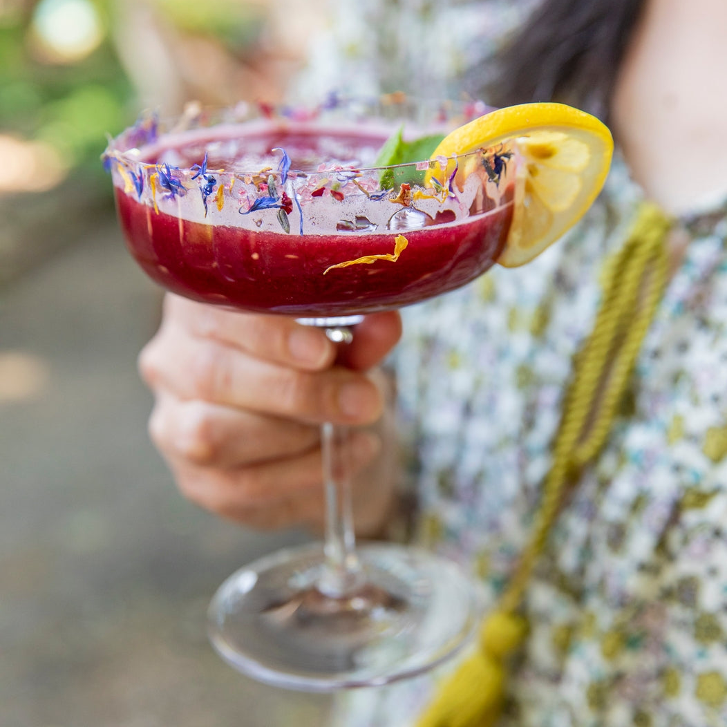 Person holding a colorful cocktail with a lemon wedge in an outdoor setting