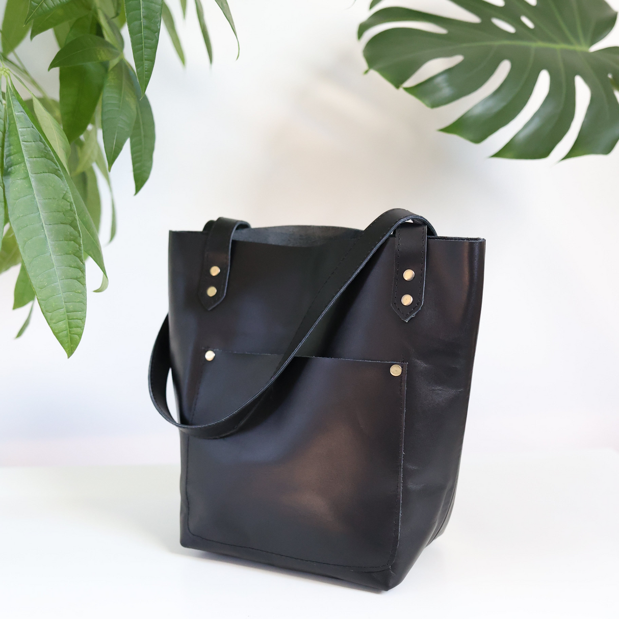 A black leather tote bag with two handles and an exterior pocket, displayed against a white background with a green plant leaf partially visible to the left.