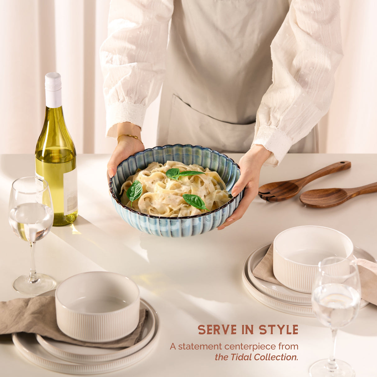 Person holding a bowl of pasta with a bottle of wine and glasses on a table, featuring the Tidal Collection.