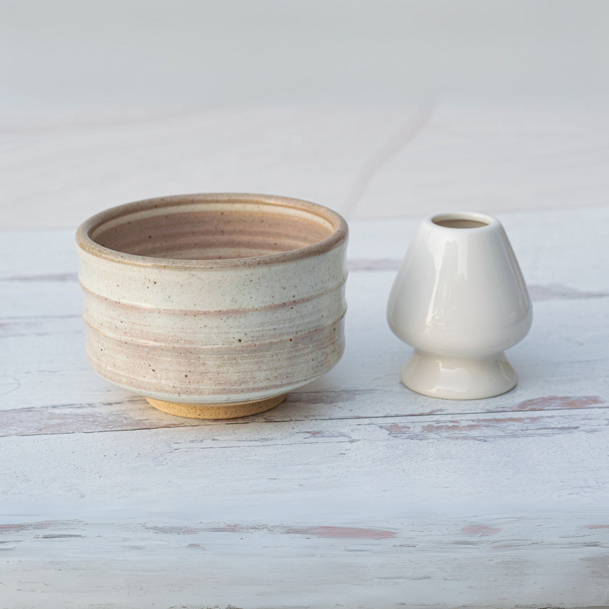 A Beige Ceramic Matcha Bowl from the Bamboo Matcha Whisk, Whisk Holder, and Organic Japanese Matcha Set rests gracefully alongside a small white vase on a light wooden surface.