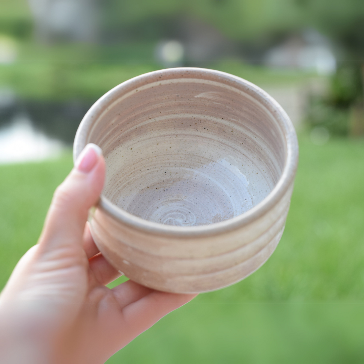 A hand holds a handmade bowl reminiscent of Japanese pottery. The image's blurry greenery enhances the serene feel. Ideal for tea lovers, this Beige Ceramic Matcha Bowl, part of a set with a Bamboo Whisk and Organic Matcha, makes an excellent matcha accessory.