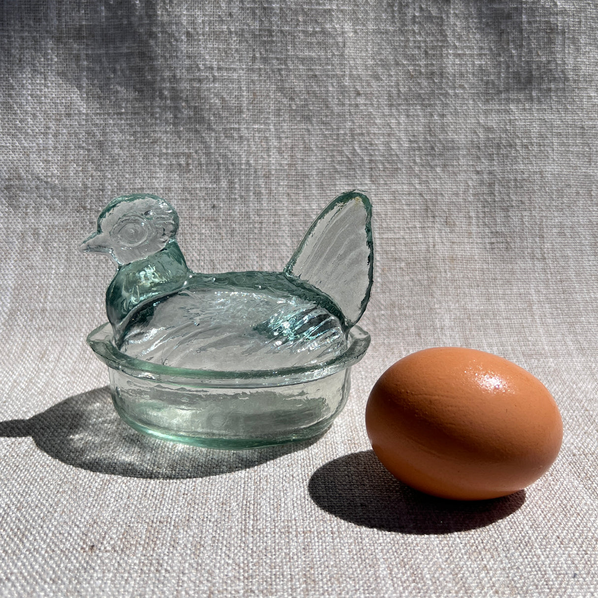 Glass hen-shaped container with a lid next to a brown egg on a textured fabric background