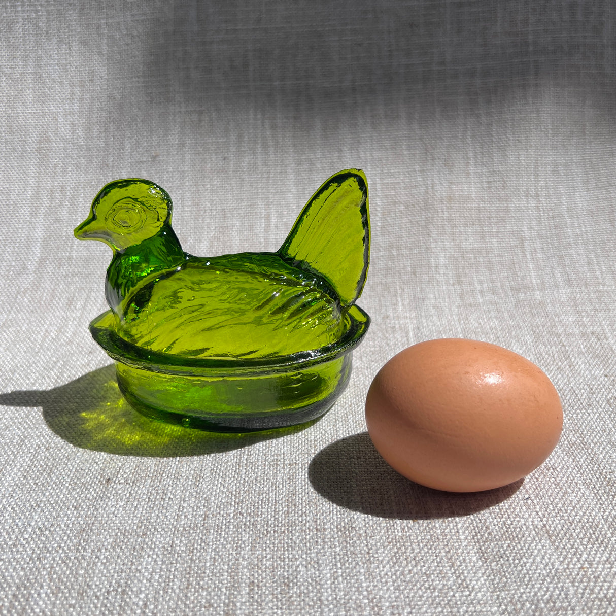 Green glass nesting hen-shaped bowl & lid next to a brown egg on a textured fabric background