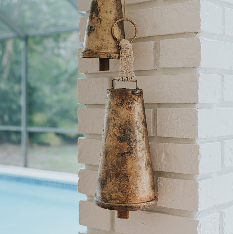 Rustic metal bell attached to a white brick wall with a pool and greenery in the background.