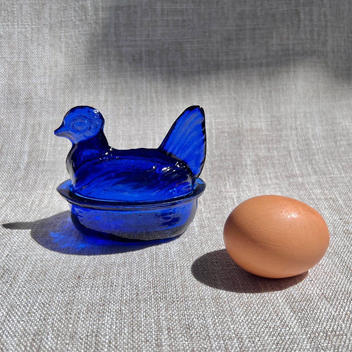 Blue glass hen-shaped bowl with a brown egg on a textured fabric background