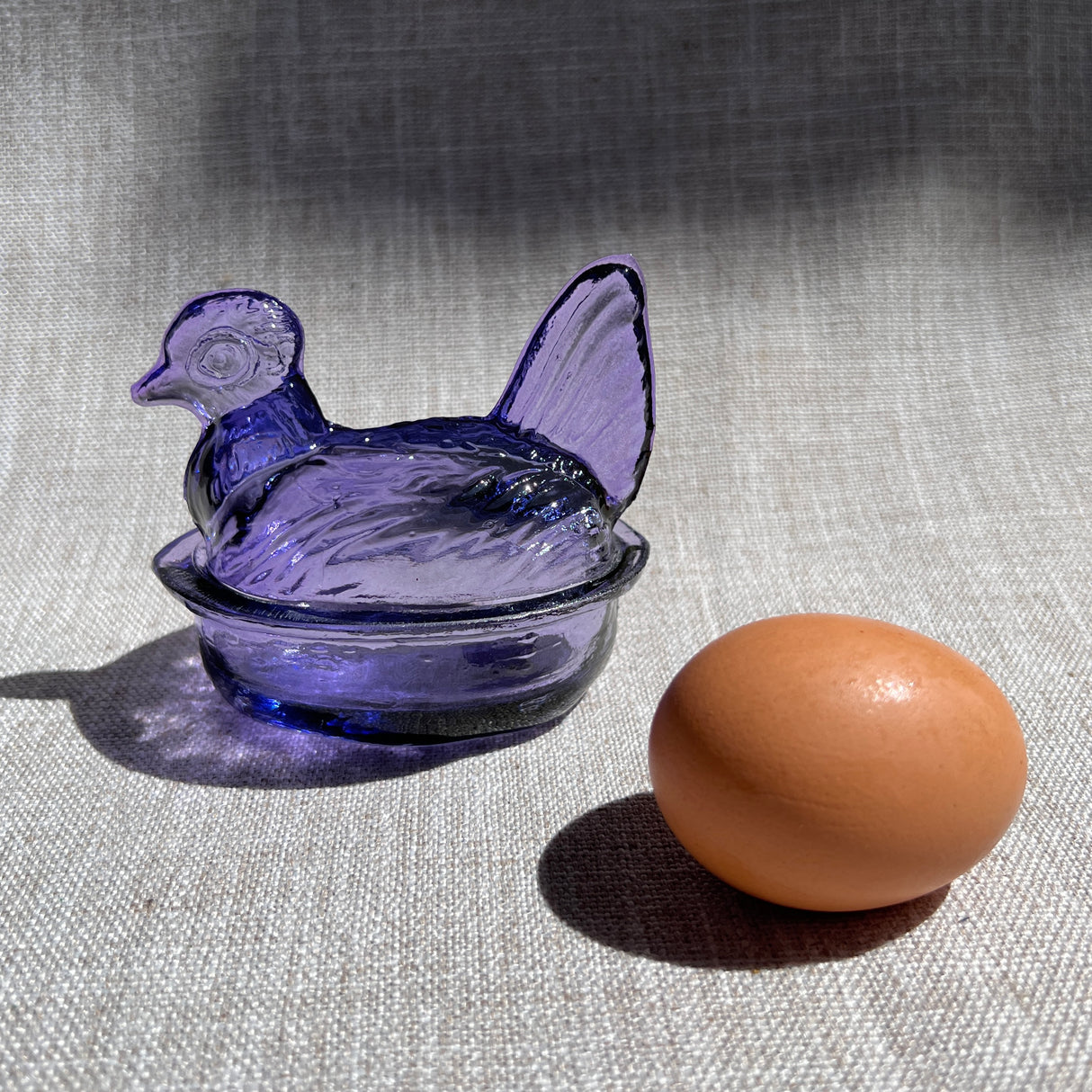 lavender glass hen-shaped bowl & lid next to a brown egg on a textured surface