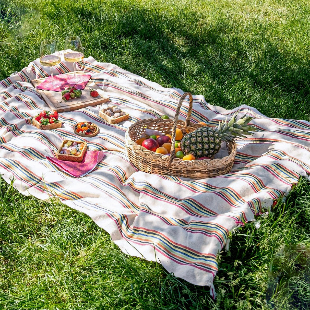 Picnic setup on a striped blanket with a basket of fruits and vegetables on a grassy field.