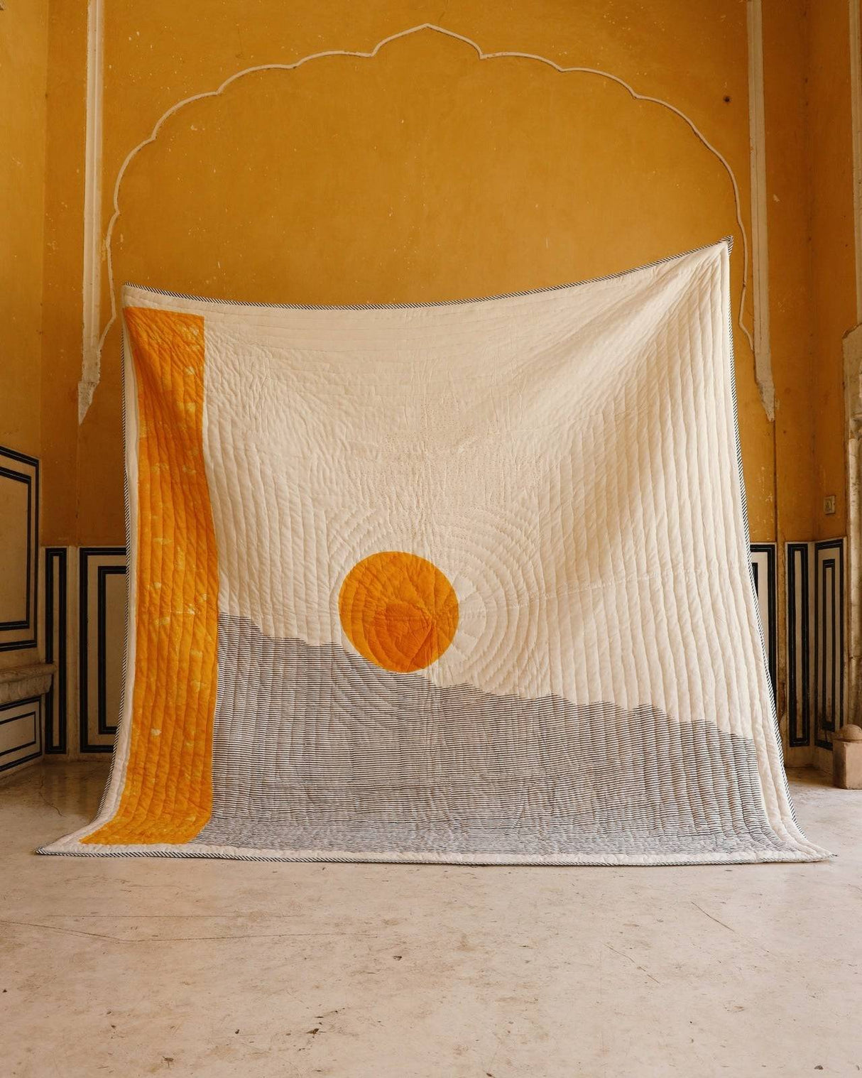 Textile art piece with a sun design on a yellow wall.