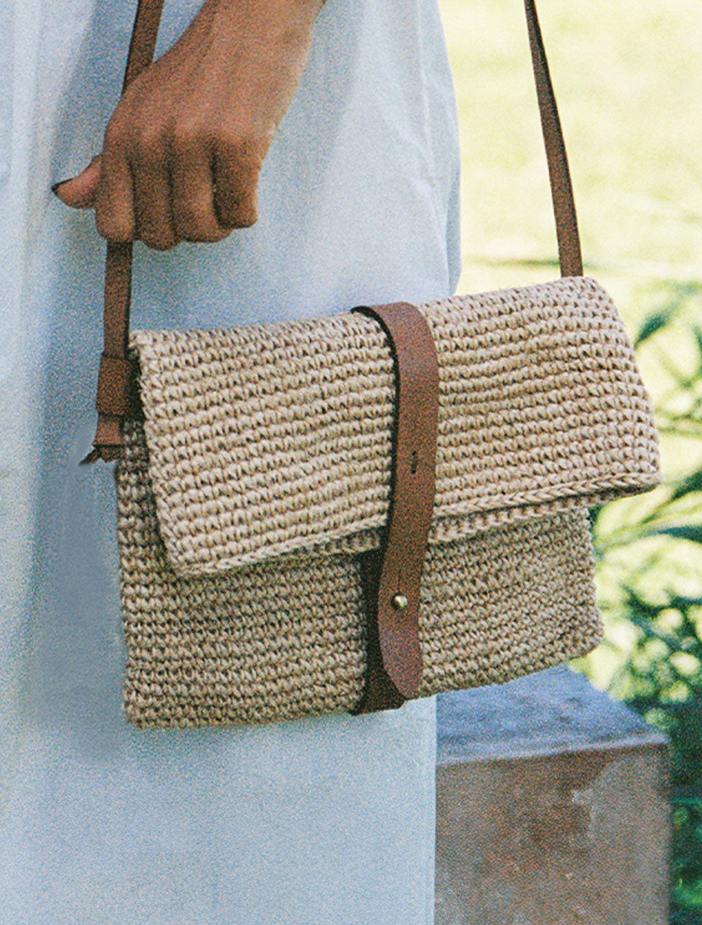 Woven handbag with brown strap held by a person against a blurred natural background