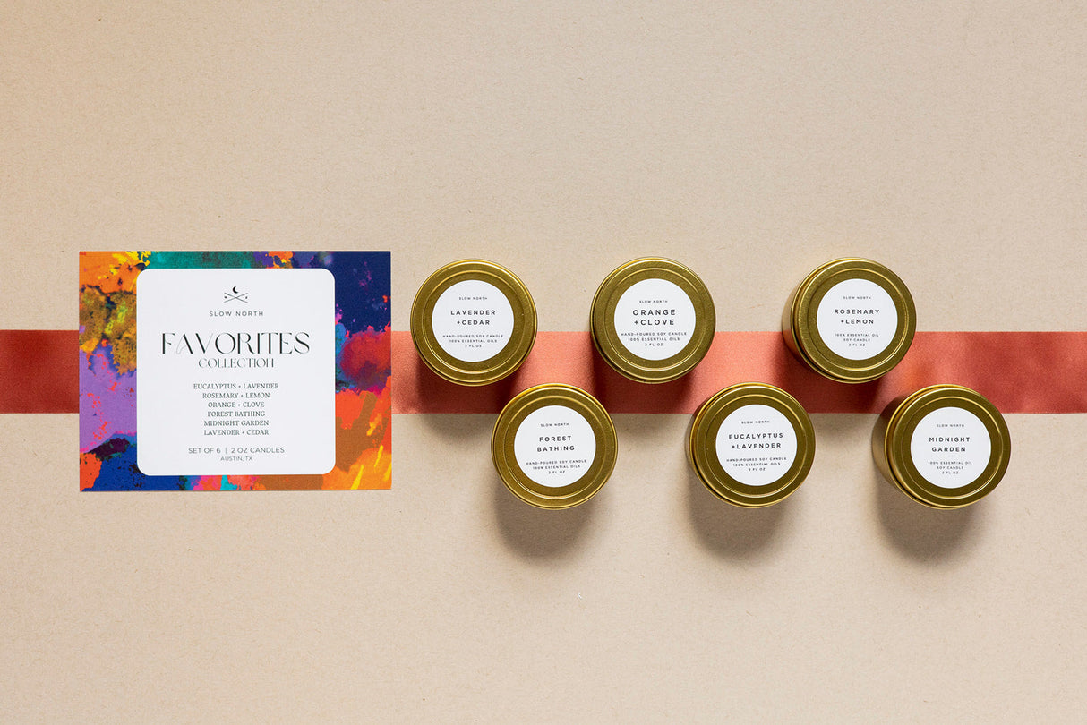 Set of six small candles with labels on a beige surface, accompanied by a colorful card. 2oz Mini Tins 6 Count