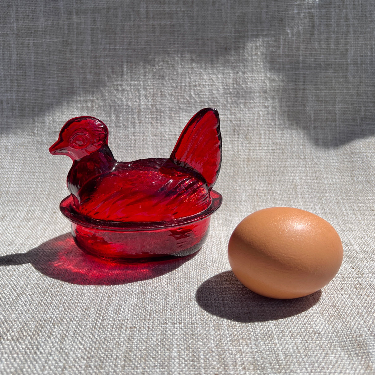 Red glass nesting hen-shaped bowl & lid next to a brown egg on a textured surface