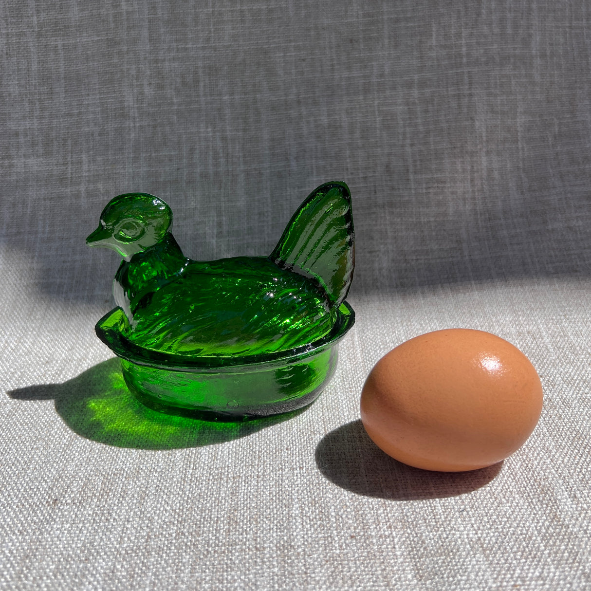 Green glass chicken-shaped container with a brown egg on a gray fabric background