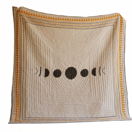 Decorative quilt with moon phase design on a wooden floor against an orange wall.