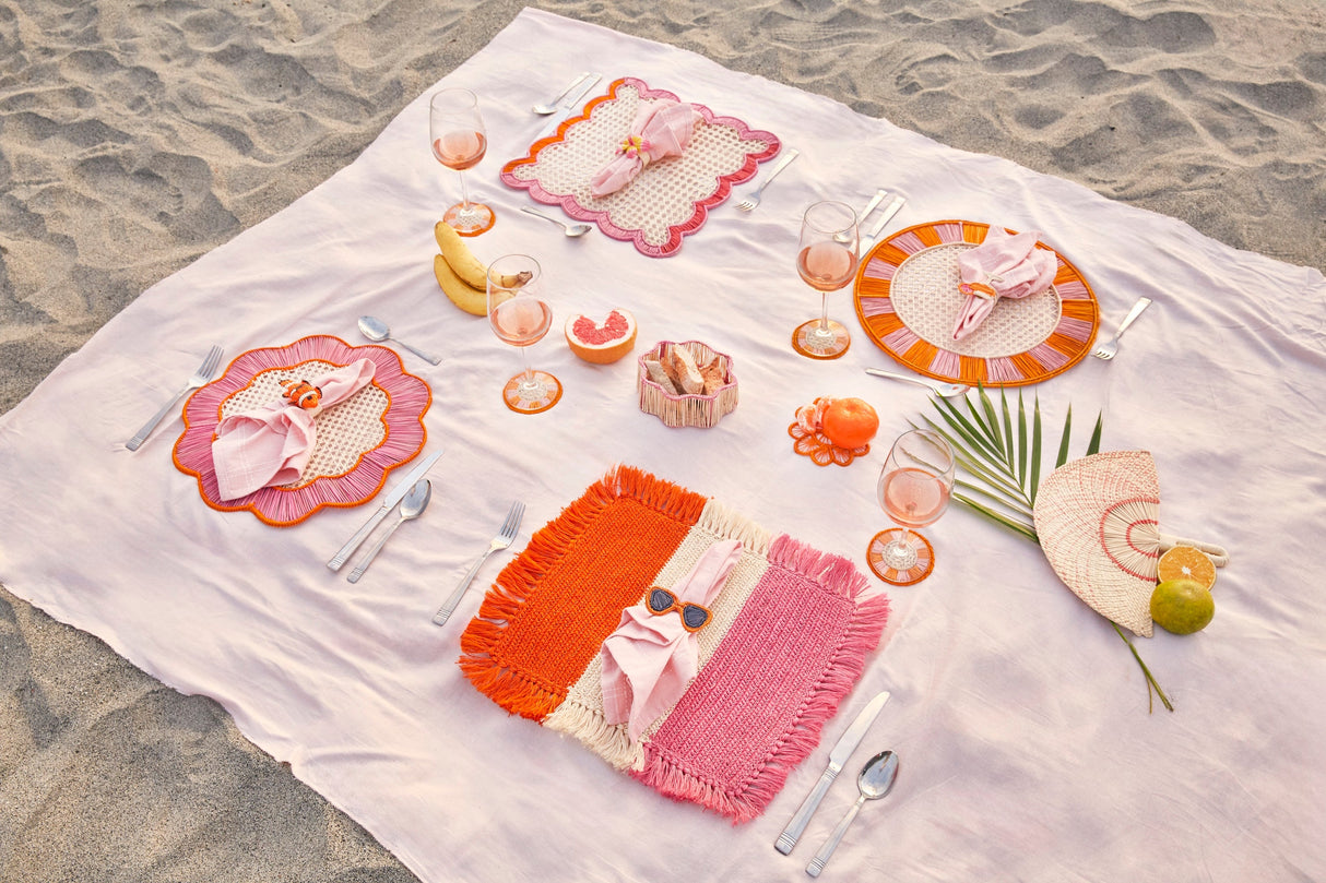 Set table with colorful plates, cutlery, and glasses on a white blanket on sand.