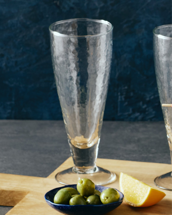 Clear glass with a bowl of olives and a lemon on a wooden surface against a dark background