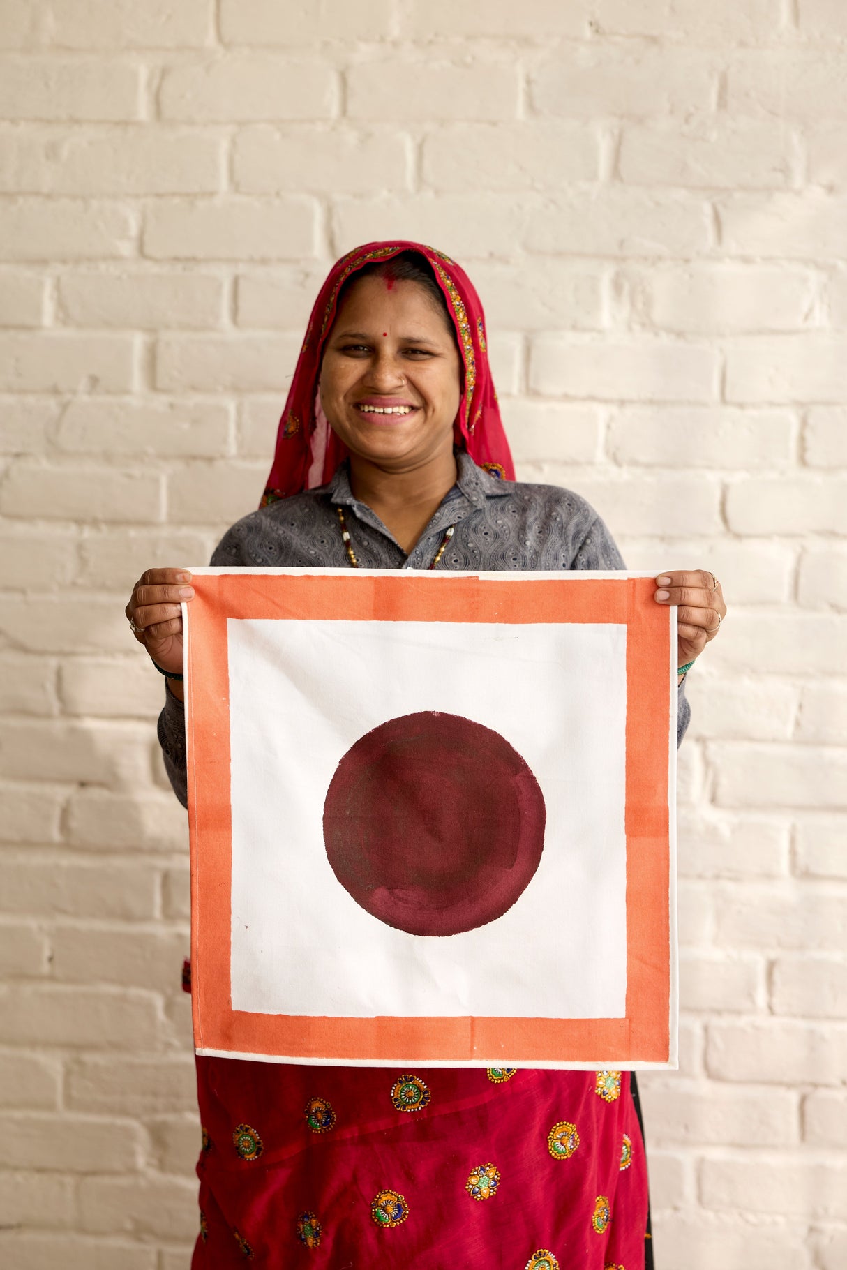 Woman holding a framed artwork with a red circle on a white background against a brick wall.