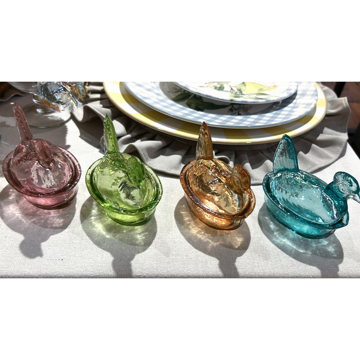 Four colorful glass hen figurines on a table setting with plates and napkins.