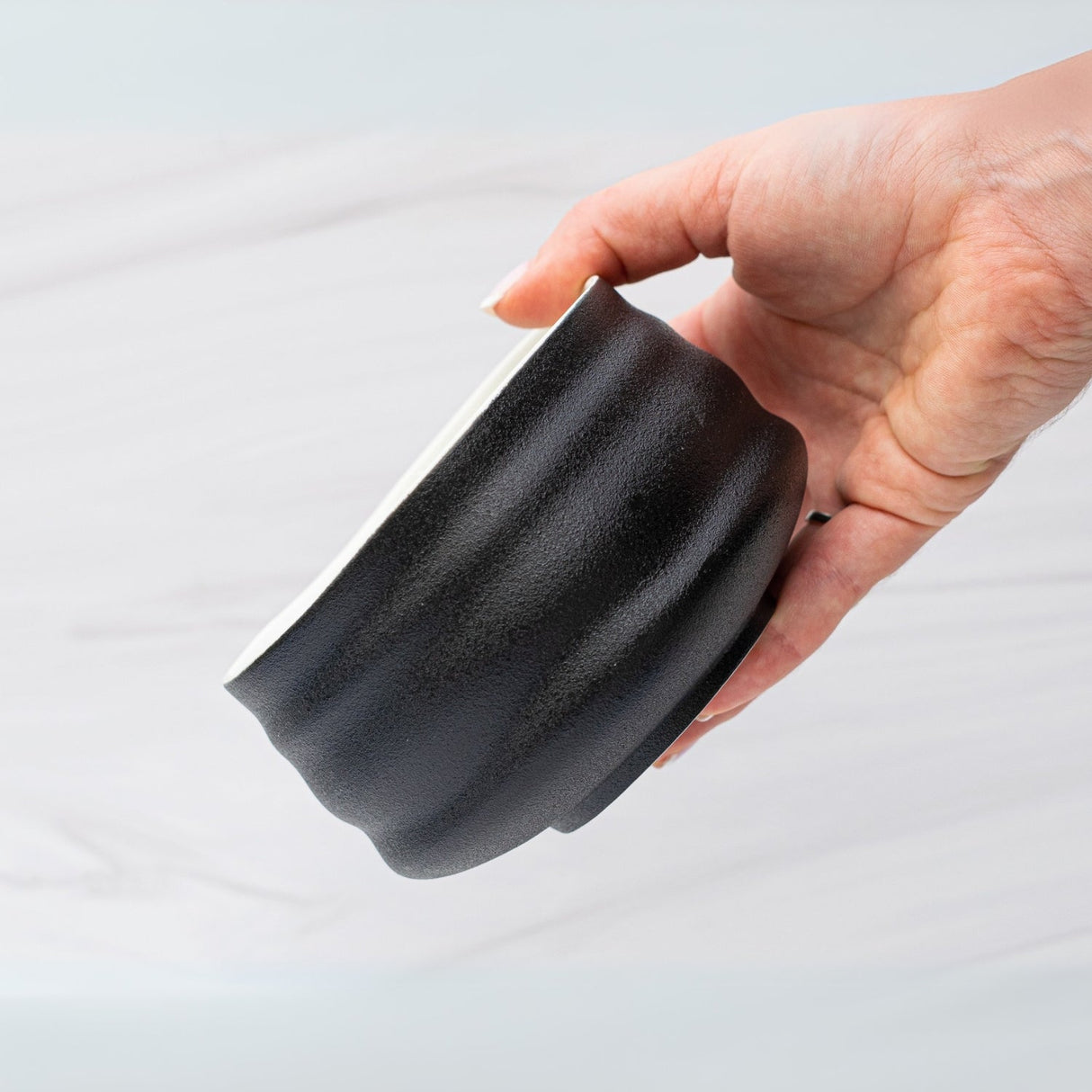 A hand gracefully holds a Black and White Ceramic Matcha Bowl, featuring a textured wave pattern, against a light background.
