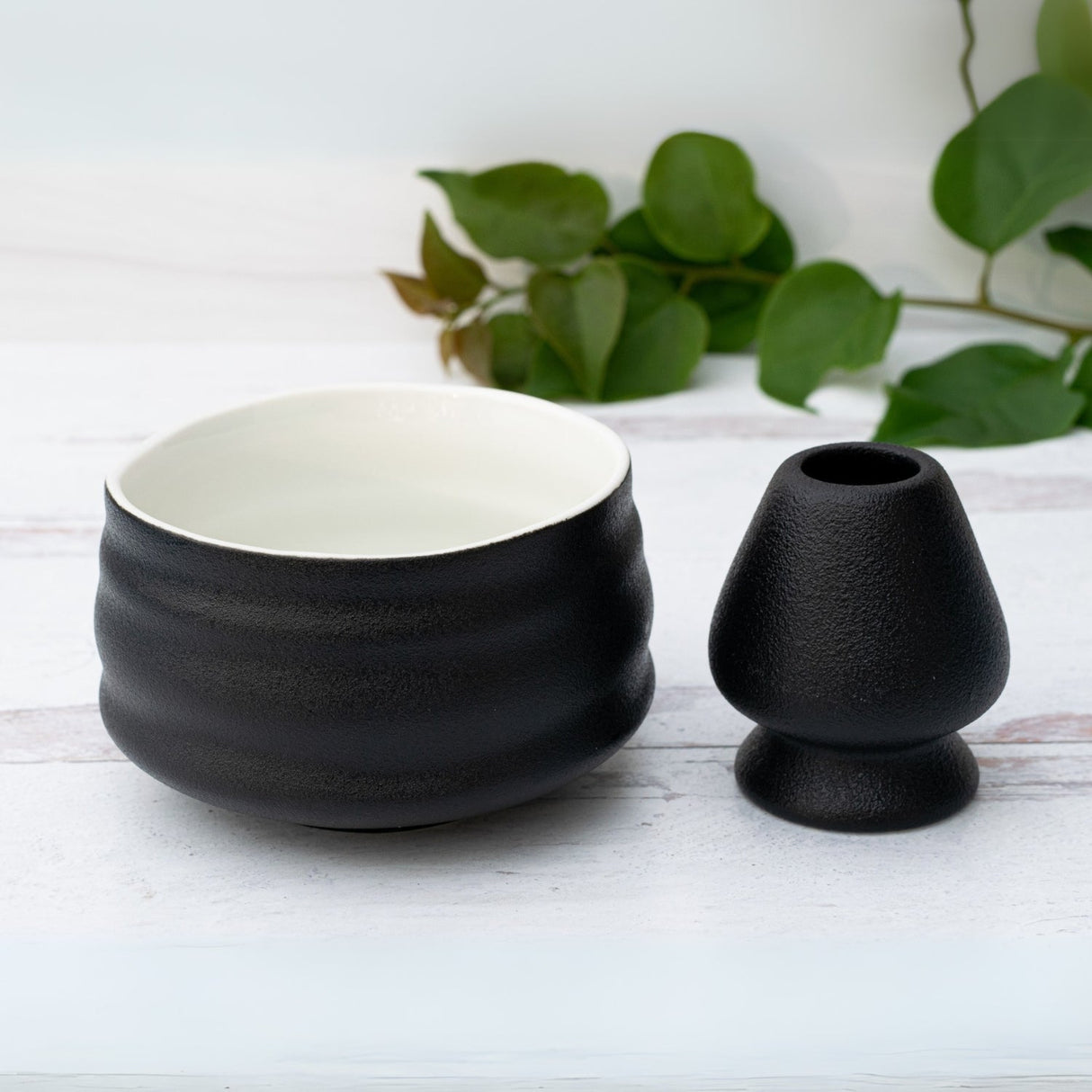 A Black and White Ceramic Matcha Bowl, evoking traditional Minoyaki pottery, rests gracefully on a white wooden surface with green leaves. Paired perfectly with a bamboo matcha whisk, it creates an exquisite matcha tea set.