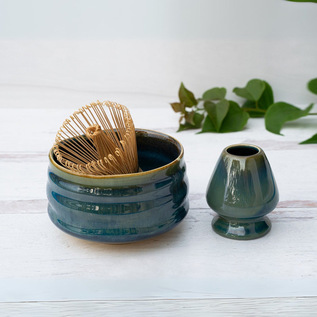 A Blue Ceramic Matcha Bowl and Bamboo Matcha Whisk from the Organic Japanese Matcha Set rests on a white surface with a small vase, surrounded by green leaves, highlighting Minoyaki pottery's serene beauty.
