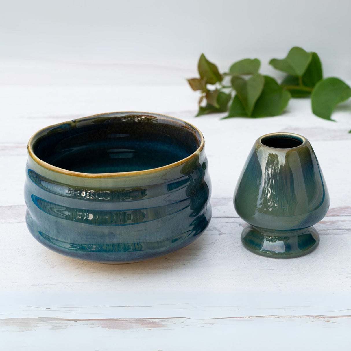 The Blue Ceramic Matcha Set, featuring a bamboo whisk, whisk holder, and organic Japanese matcha, is arranged on a white surface with green leaves in the background.