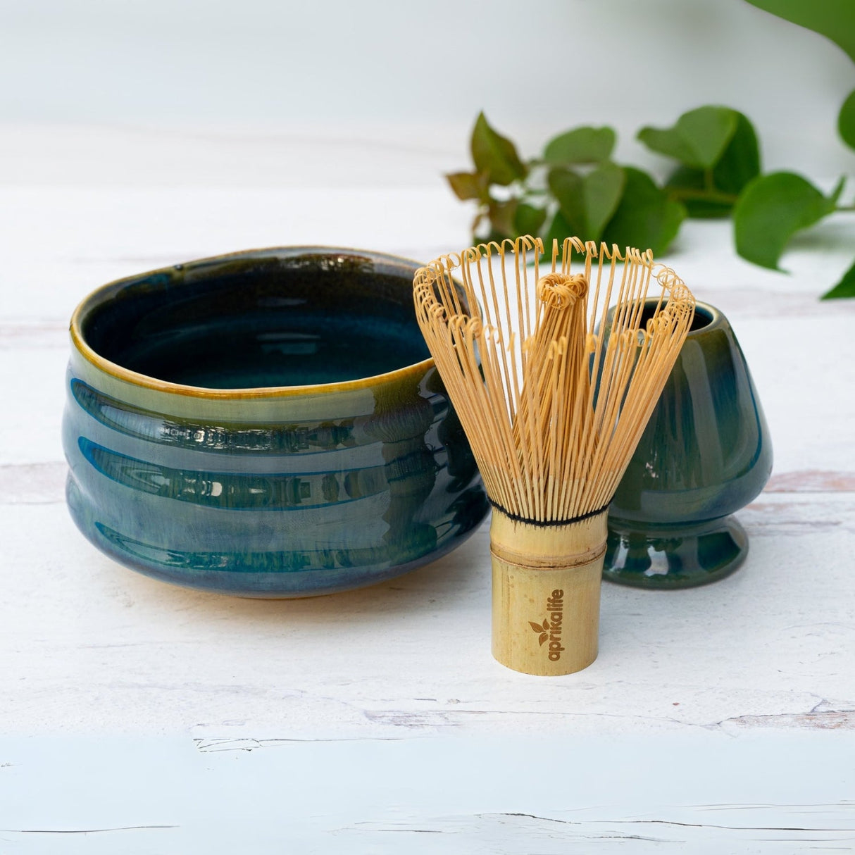 On a white surface, a Blue Ceramic Matcha Bowl sits elegantly beside a Bamboo Matcha Whisk from the Organic Japanese Matcha Set, framed by lush green leaves.