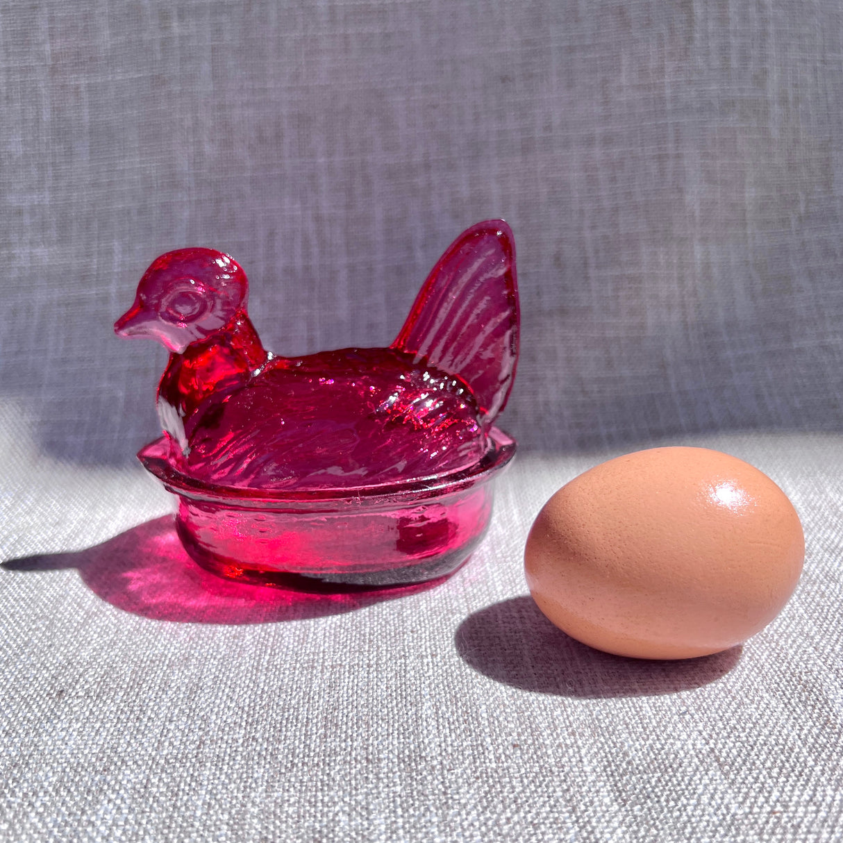 Magenta glass chicken-shaped egg holder next to a brown egg on a textured surface
