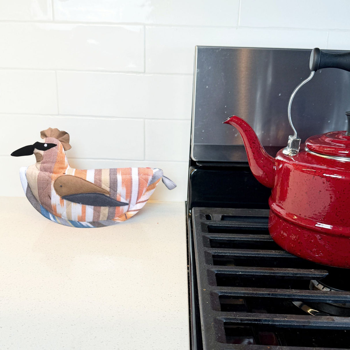 Red kettle on a stove with a decorative towel and duck-shaped object in the background.