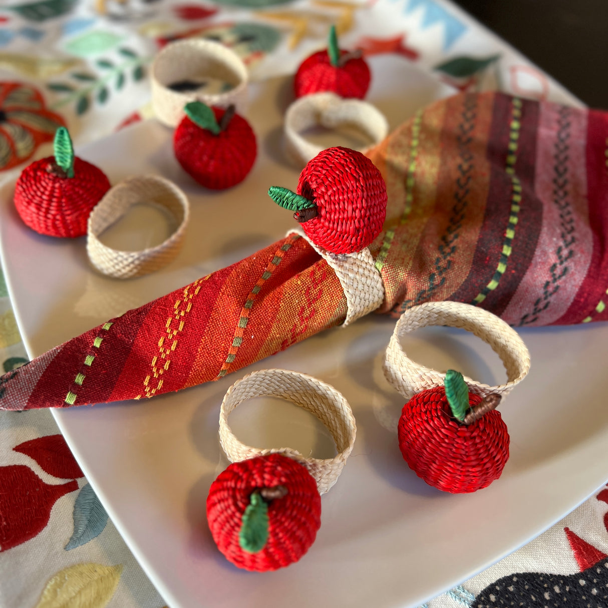 Decorative napkin rings shaped like red apples on a white plate with a colorful tablecloth.