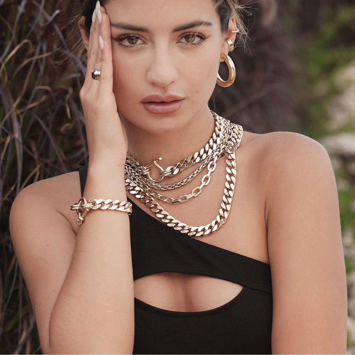Woman wearing gold jewelry including necklaces, earrings, and bracelet against a blurred natural background