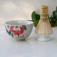 The Red and Blue Flowers Matcha Bowl, Bamboo Matcha Whisk, and Whisk Holder Set is showcased on a light surface with green leaves in the background, highlighting Minoyaki pottery techniques.