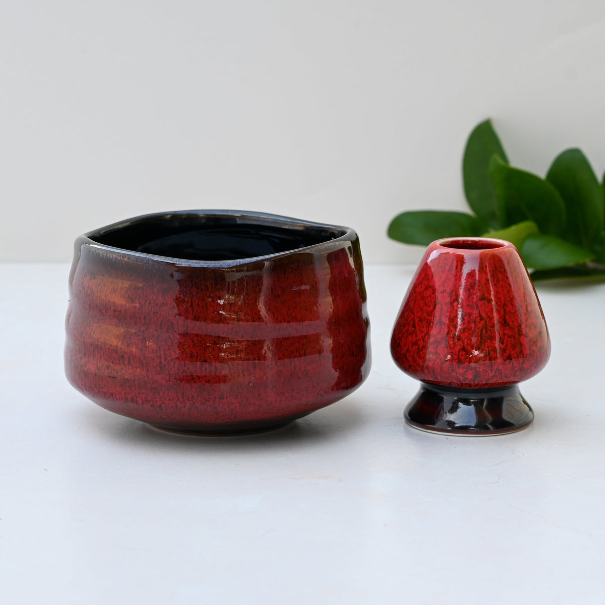 A Red Matcha Bowl with a glossy finish sits elegantly on a light surface, reminiscent of Minoyaki Pottery. Nearby, a green plant adds serenity, evoking the tranquility of traditional Matcha Tea Ceremonies.