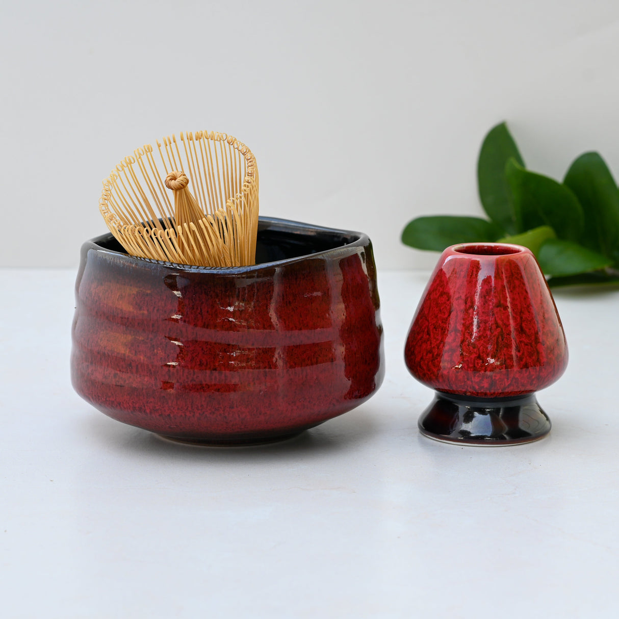 The Red Matcha Bowl, part of a Minoyaki Pottery set, includes a delicate bamboo whisk and a small pot. Vibrant green leaves sit gracefully on a pristine white surface—ideal for elegant matcha tea ceremonies.