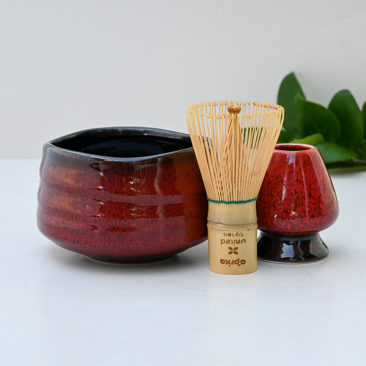 A Red Matcha Bowl, crafted with exquisite Minoyaki pottery and accompanied by a matcha whisk, is displayed elegantly on a white surface with green leaves in the background.