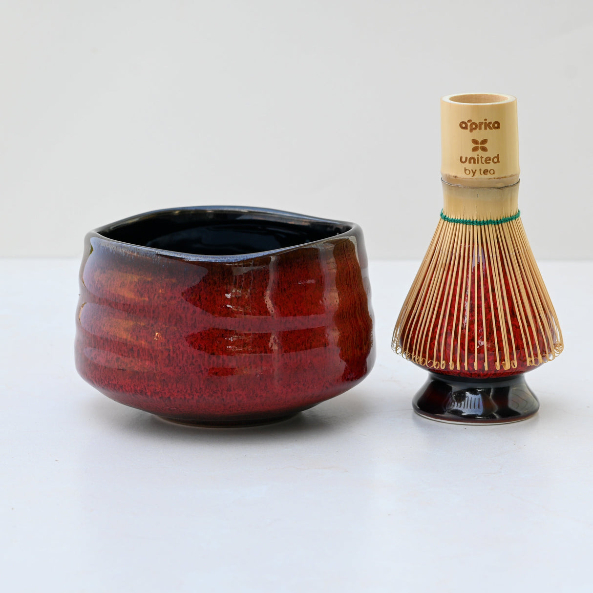 A Red Matcha Bowl sits beside a bamboo tea whisk with colorful bristles on a light surface, perfect for traditional matcha tea ceremonies.