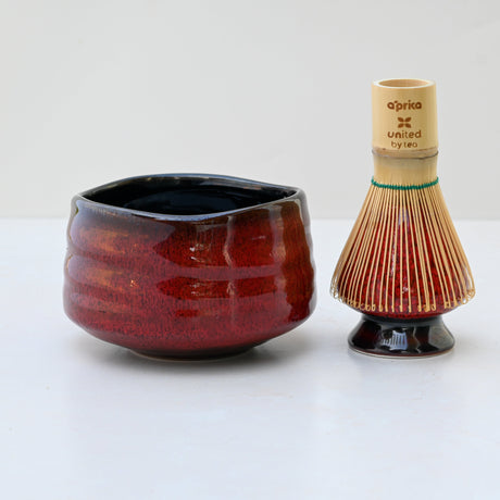 A Red Matcha Bowl sits beside a bamboo tea whisk with colorful bristles on a light surface, perfect for traditional matcha tea ceremonies.