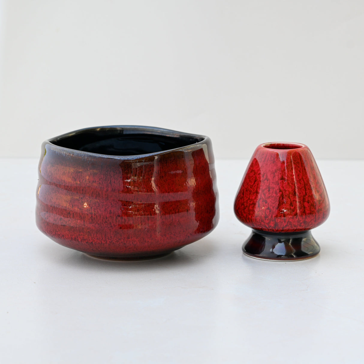 A Red Matcha Bowl, crafted with Minoyaki pottery techniques, features a glossy, textured finish and sits on a white surface alongside a ceramic vase.