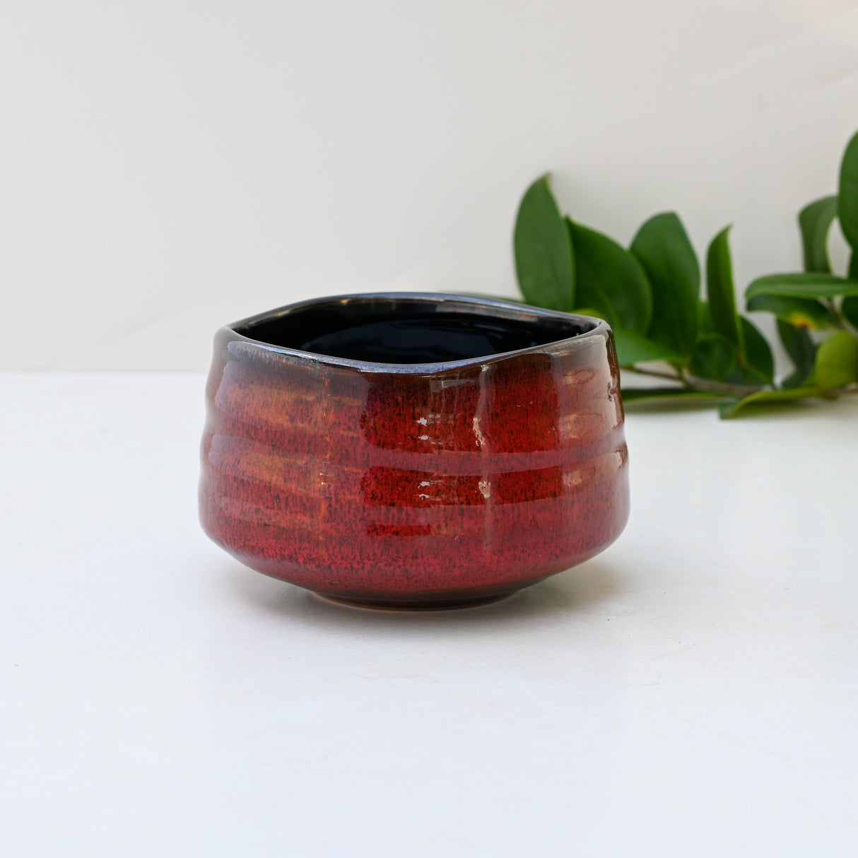 A Red Matcha Bowl, crafted in the Minoyaki style, sits on a white background, surrounded by green leaves.