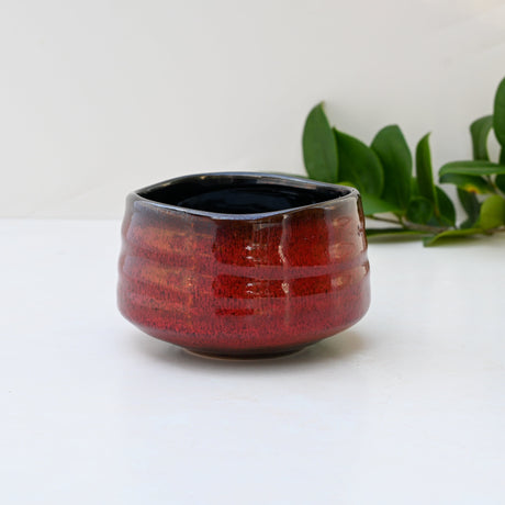 A Red Matcha Bowl, crafted in the Minoyaki style, sits on a white background, surrounded by green leaves.