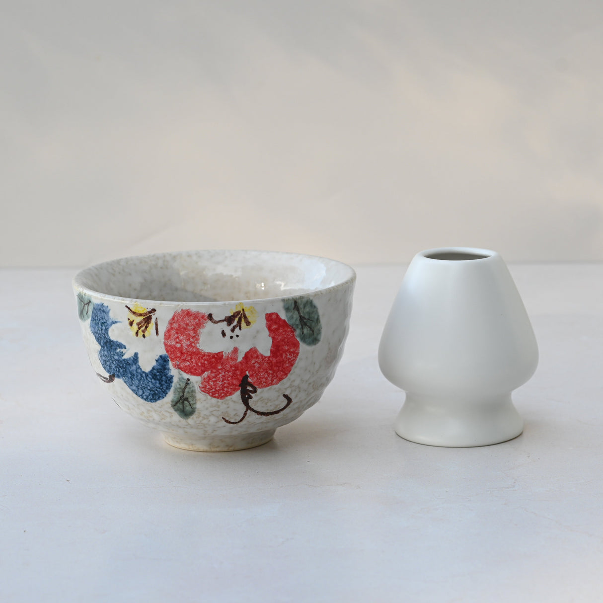 A Red and Blue Flowers Matcha Bowl, crafted with Minoyaki pottery techniques, displays colorful floral designs alongside a small white vase on a light surface.