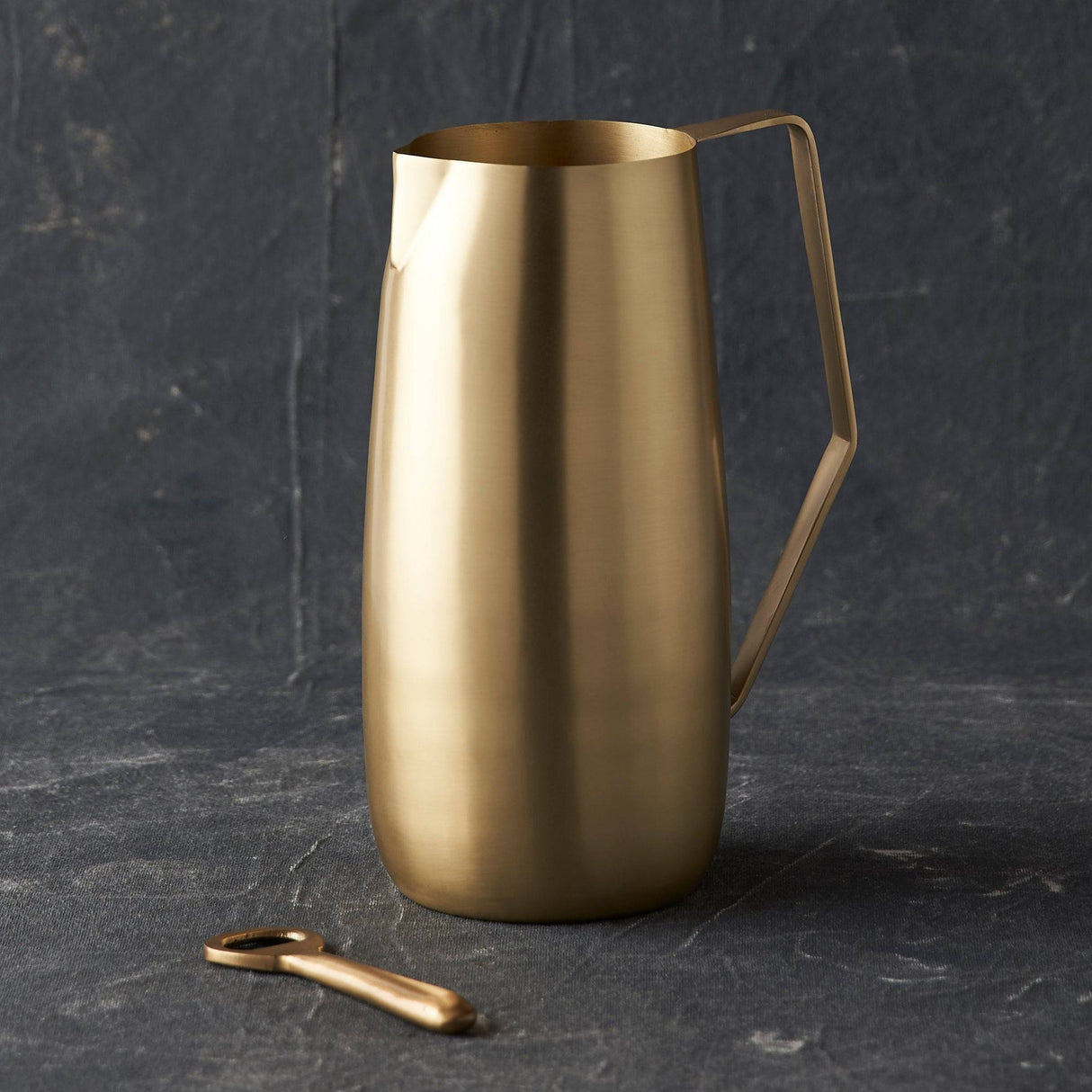 A gold-colored brass pitcher with an angled handle, displayed against a dark background.