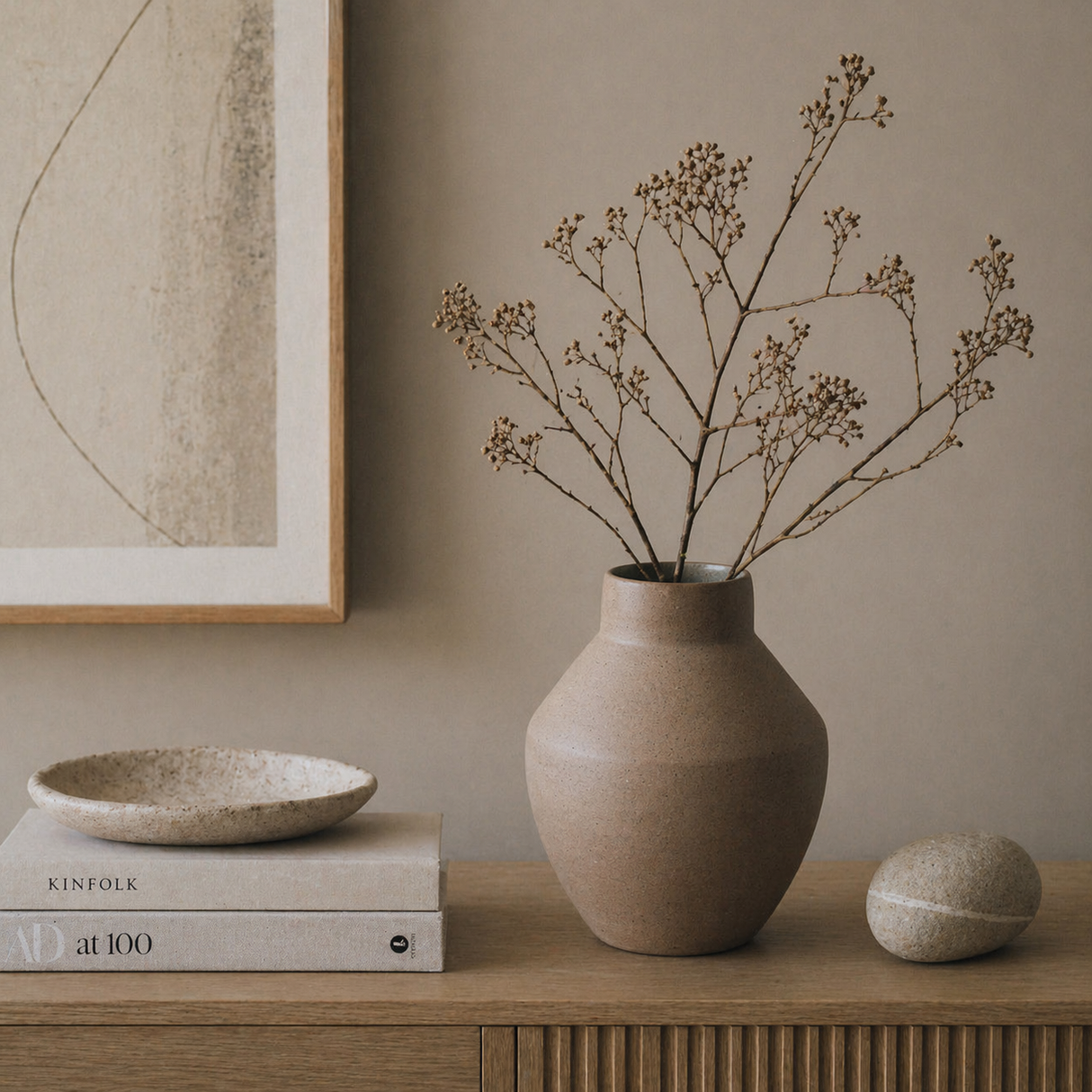 Ceramic vase with dried branches on a wooden surface, with books and a bowl in the background.