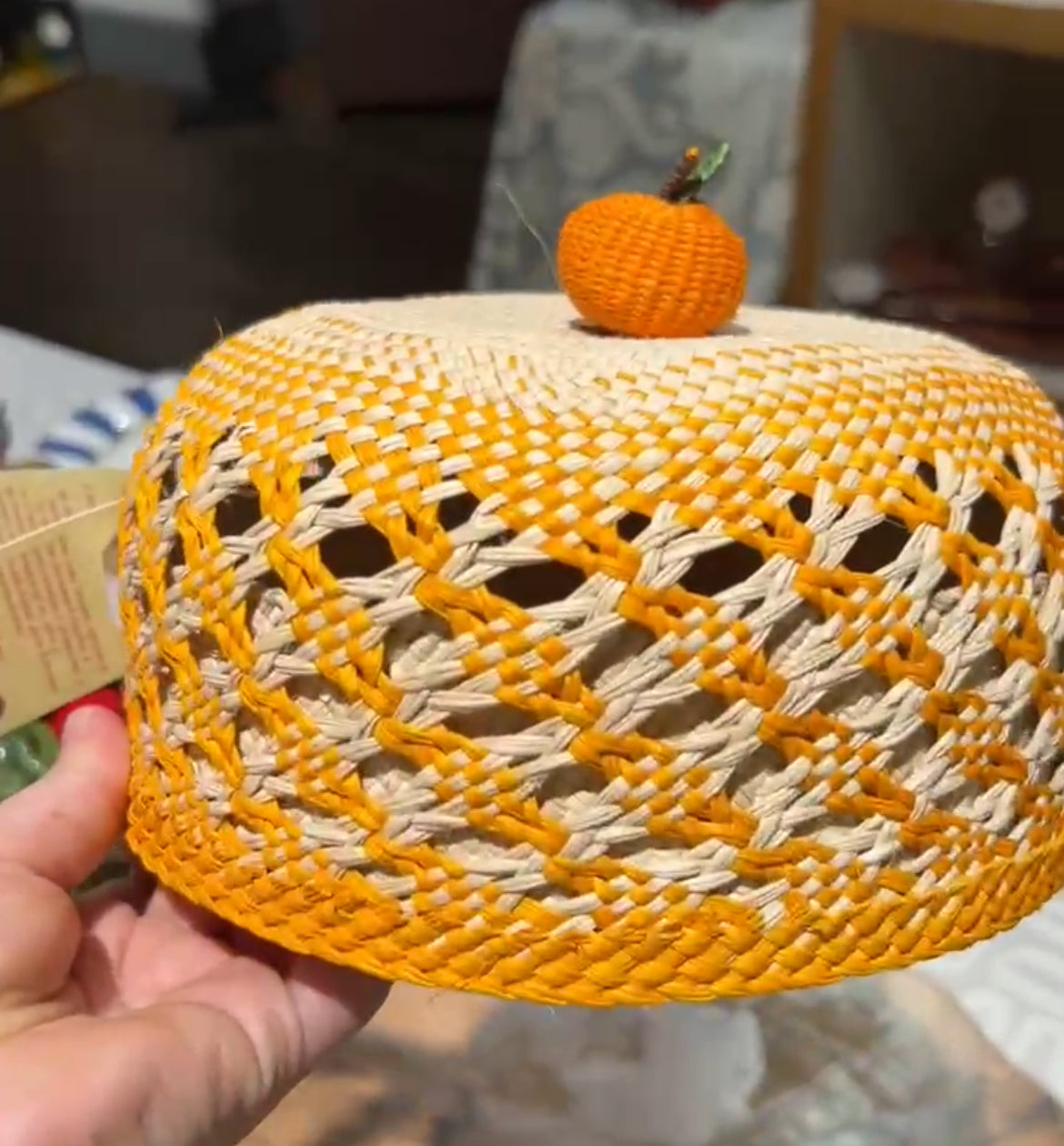 Hand holding a woven orange and beige basket lid with a small pumpkin decoration.
