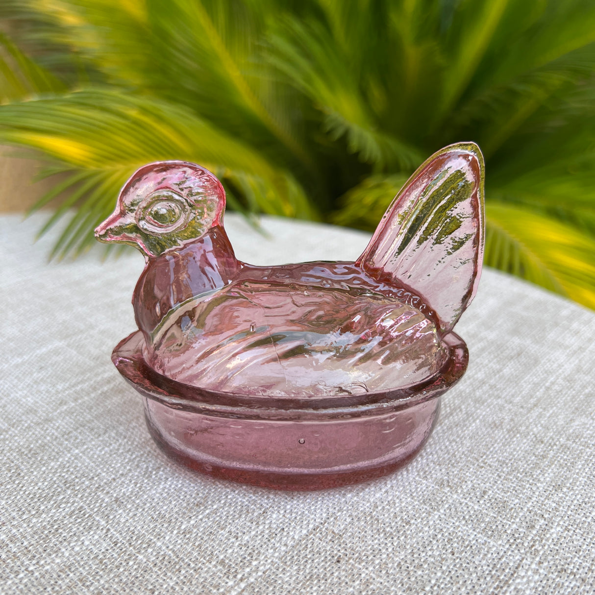 Pink glass hen-shaped bowl on a textured surface with green foliage in the background