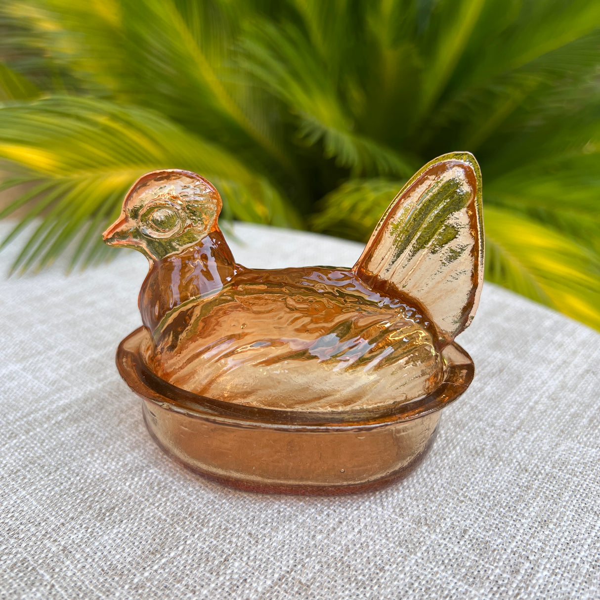 Glass chicken-shaped container on a textured surface with green foliage in the background