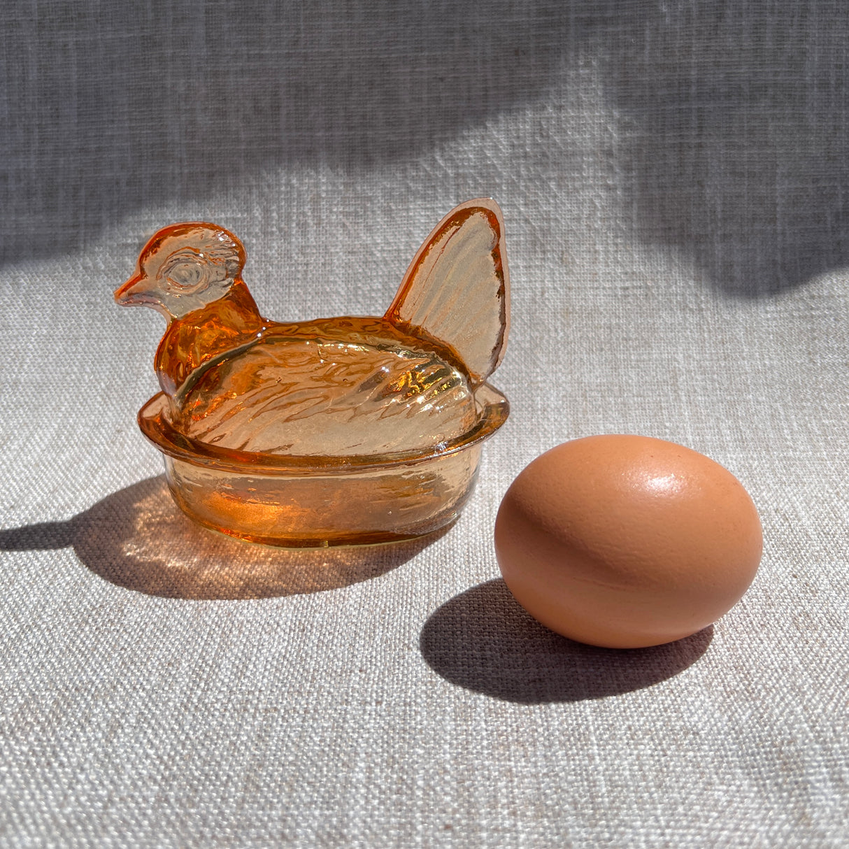 Glass hen-shaped container with a lid next to a brown egg on a textured surface