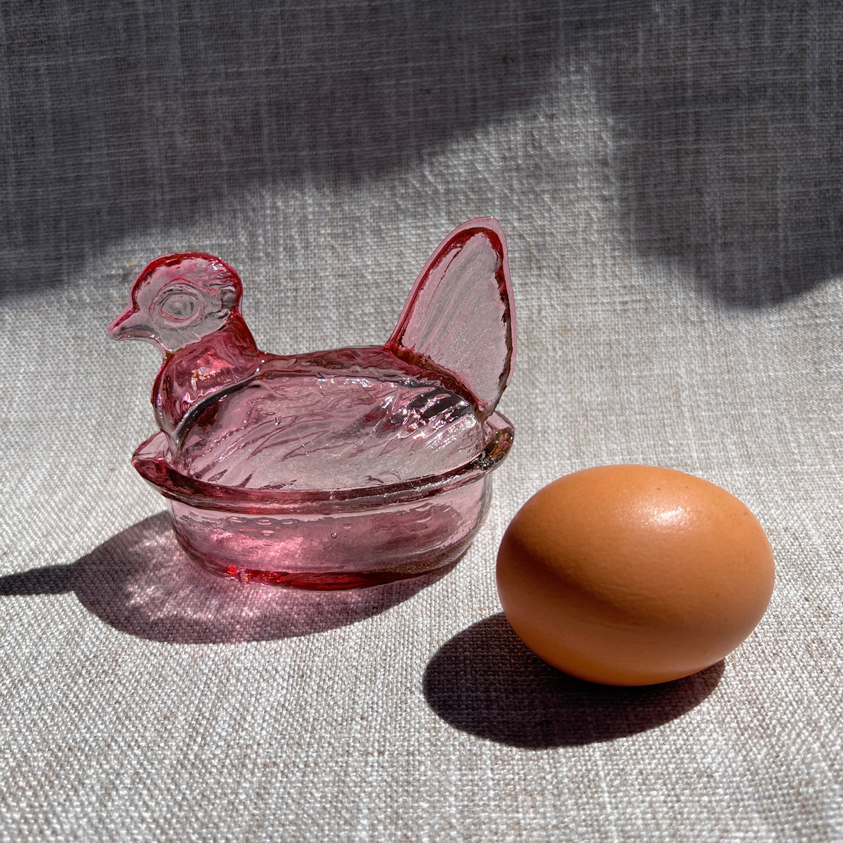 Pink glass chicken-shaped bowl next to a brown egg on a textured surface