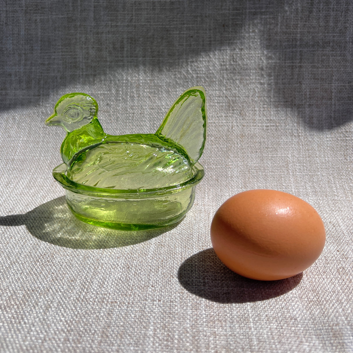 Green glass hen-shaped glass bowl next to a brown egg on a textured surface