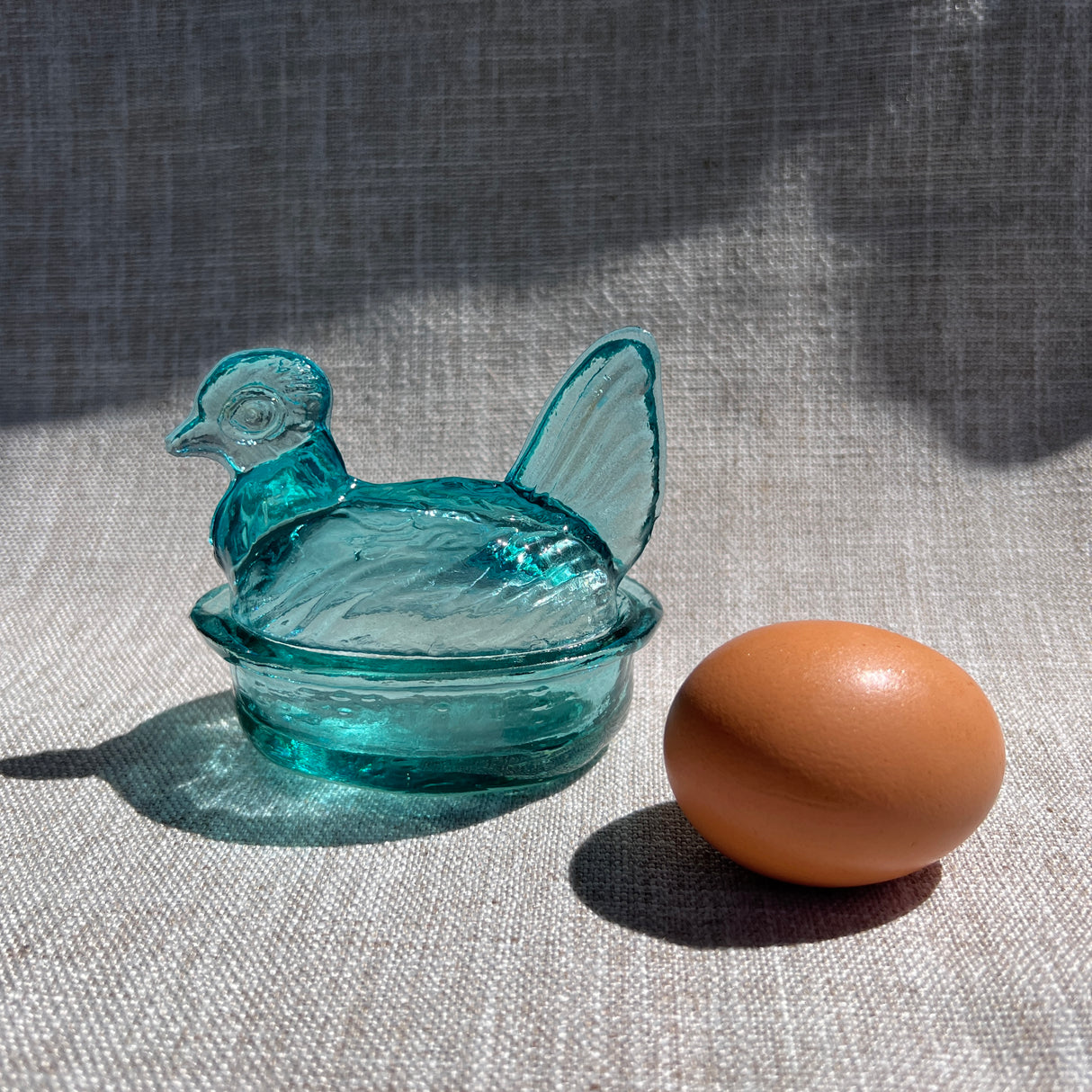 Blue glass chicken-shaped container next to a brown egg on a textured surface