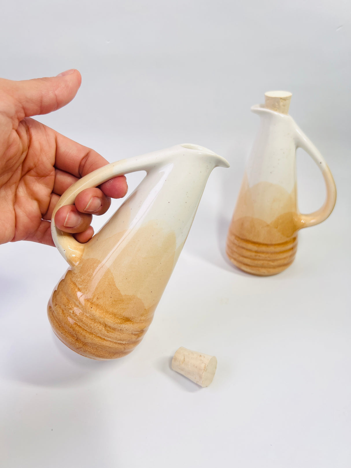 Hand holding a gradient ceramic pitcher with another pitcher in the background on a white surface.
